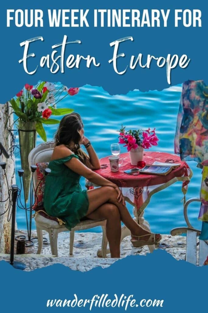Photo with text overlay. Photo in the center shows a woman sitting at a small table looking out over the water. The table holds a book, a glass of white wine and a small vase of flowers. Text reads Four Week Itinerary for Eastern Europe.
