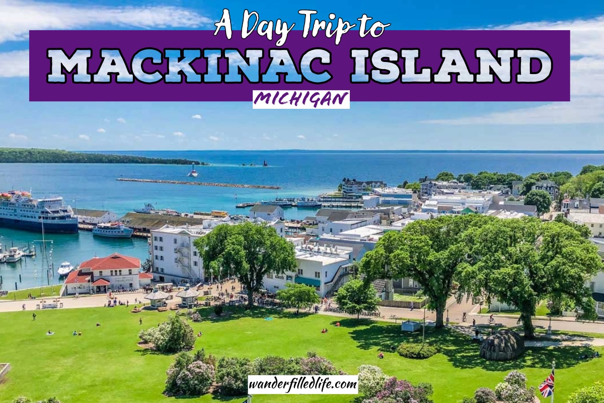 Photo with text overlay. The photo is looking out over a small downtown with a green grassy park nearby and water with a dock on the other side. Text overlay reads A Day Trip to Mackinac Island Michigan.
