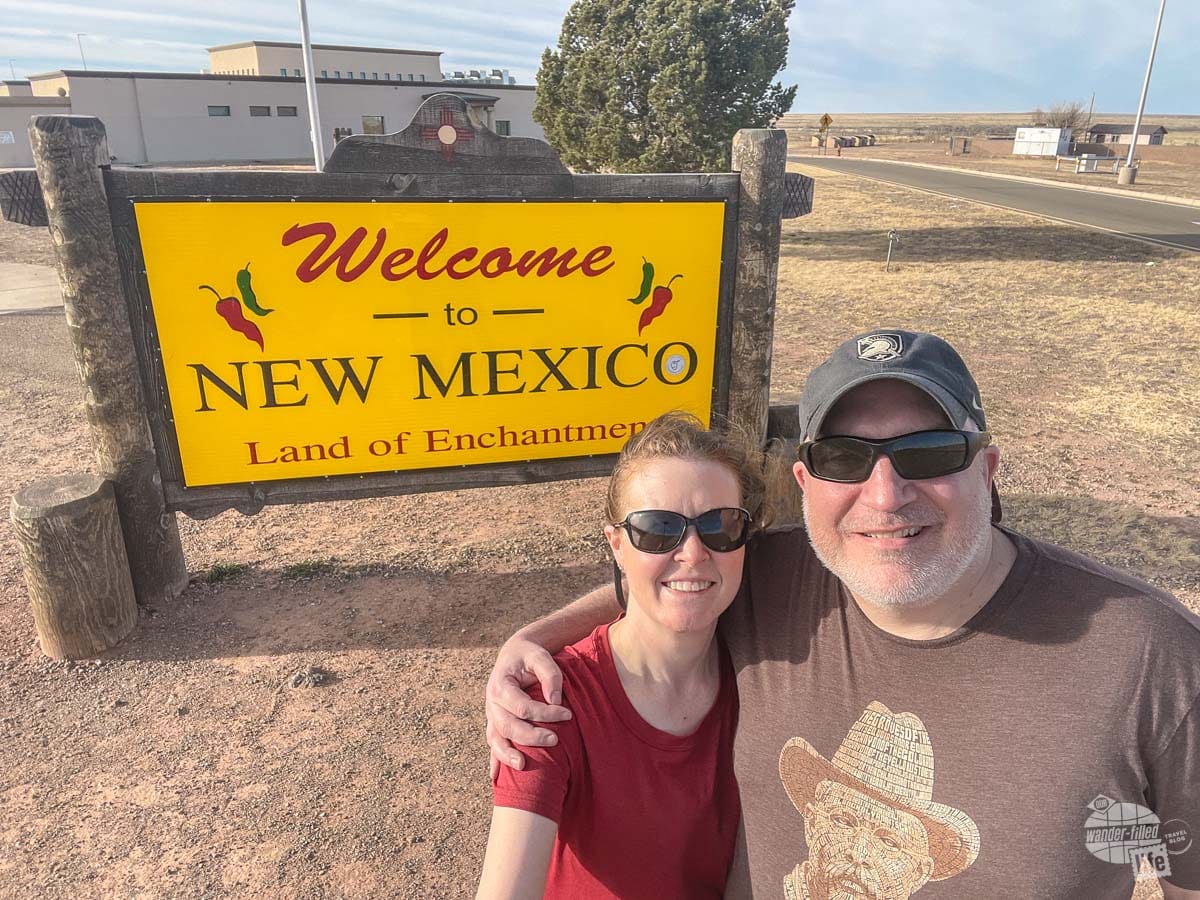 Grant and Bonnie pose in front of a yellow sign that reads "Welcome to New Mexico Land of Enchantment"