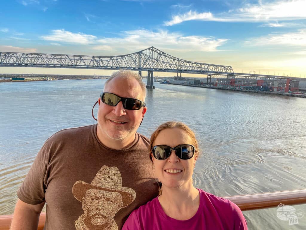 A couple pose with a steel bridge over a river in the background.