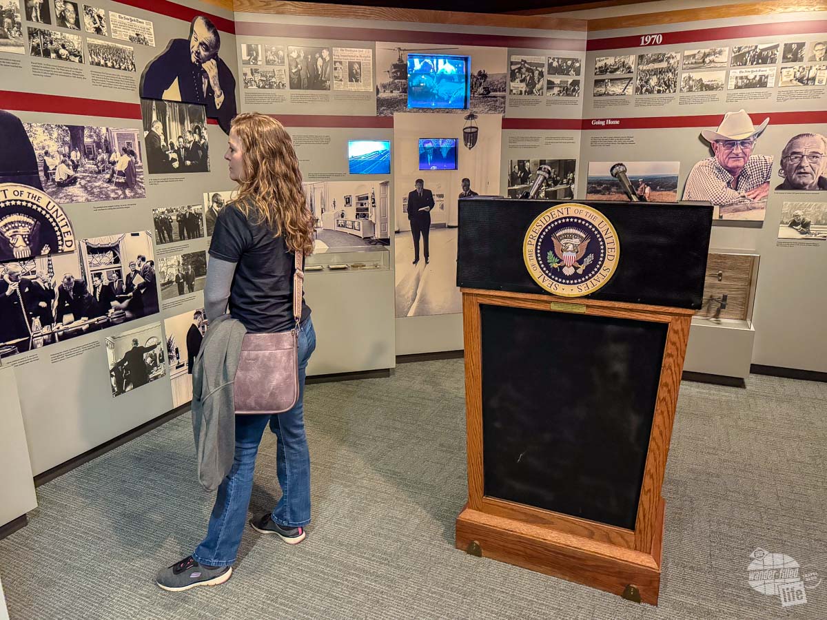 A woman standing in an exhibit with a presidential podium next to her.