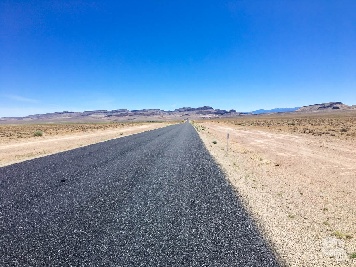 A long black stretch of road leading to the horizon with some low mountains in the distance.