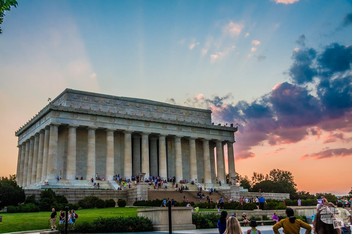 The outside of the Lincoln Memorial, surrounded by columns, with people sitting and standing all around.