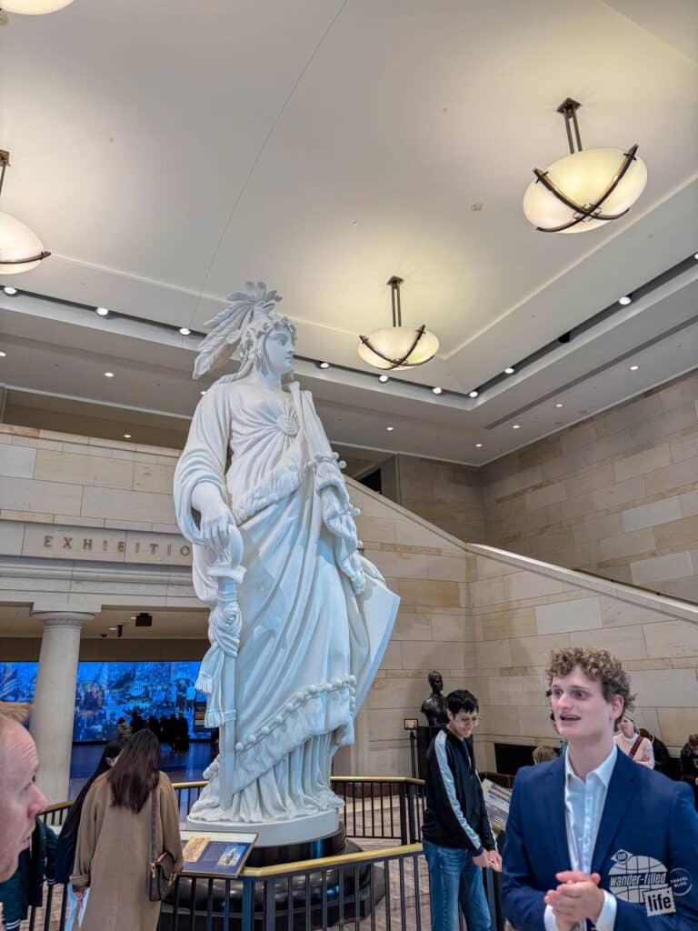 A man in a sport coat talking next to a white model of the statue of Liberty which sits atop the Capitol dome.