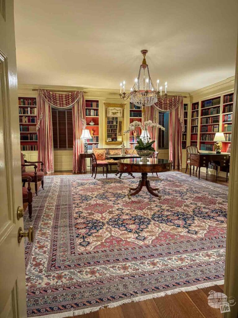 A large room with several chairs to the side and bookcases built into the wall. In the center is a mall table under a crystal chandelier.