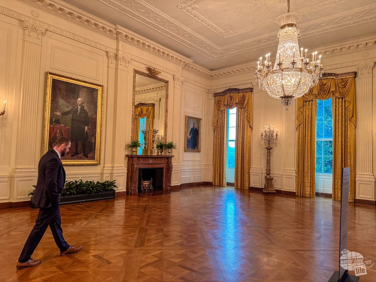 A Secret Service guard walks in a large, open room with a crystal chandelier hanging from the ceiling. There is a portrait of George Washington beside a fireplace and a portrait of Theodore Roosevelt in the corner.