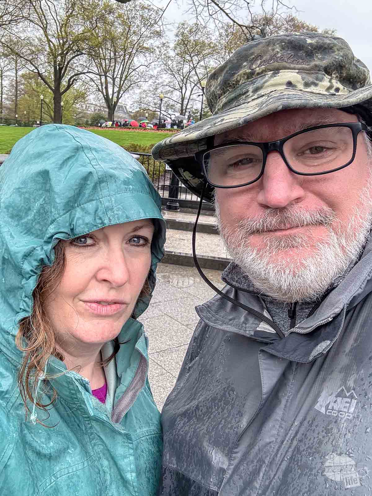 Grant and Bonnie taking a selfie in the rain. Bonnie is wearing a raincoat with a good. Grant is wearing a raincoat with a wide-brimmed camoflauge hat.