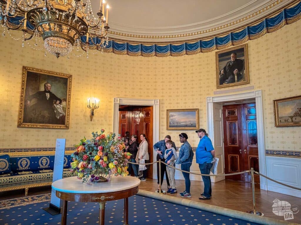 A small group of people standing outside a rope which blocks them from a blue carpet with a crystal chandelier hanging above.