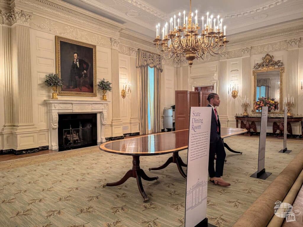 A Secret Service guard standing under a crystal chandelier in a large room with a fireplace with a portrait of Abraham Lincoln above the mantle.