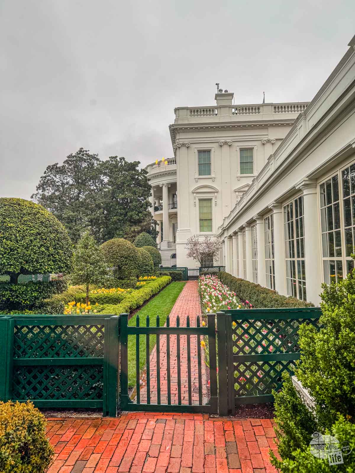 An exterior shot of the White House looking down the outside of the East Colonnade. There is a low wooden fence gating off a brick-paved walkway leading to the main building of the White House.