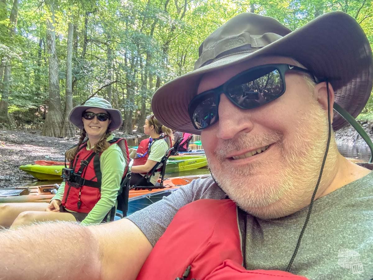 A selfie of two people with life jackets on sitting on kayaks on a creek.
