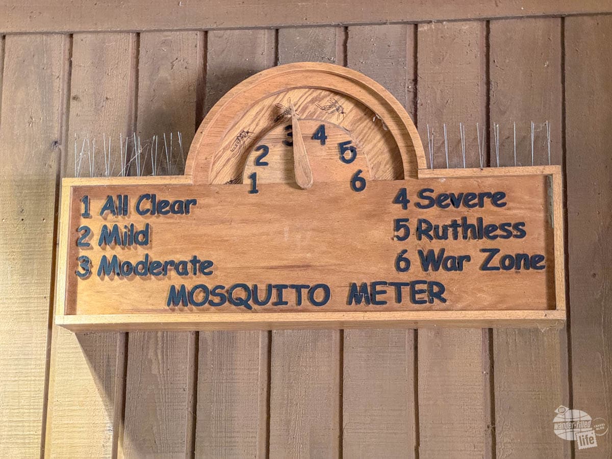 A wooden sign that says Mosquito Meter with a dial that goes from one to six. One is 'All Clear." Two is "Mild." Three is "Moderate." Four is "Severe." Five is "Ruthless." Six is "War Zone." The meter is currently set at three.