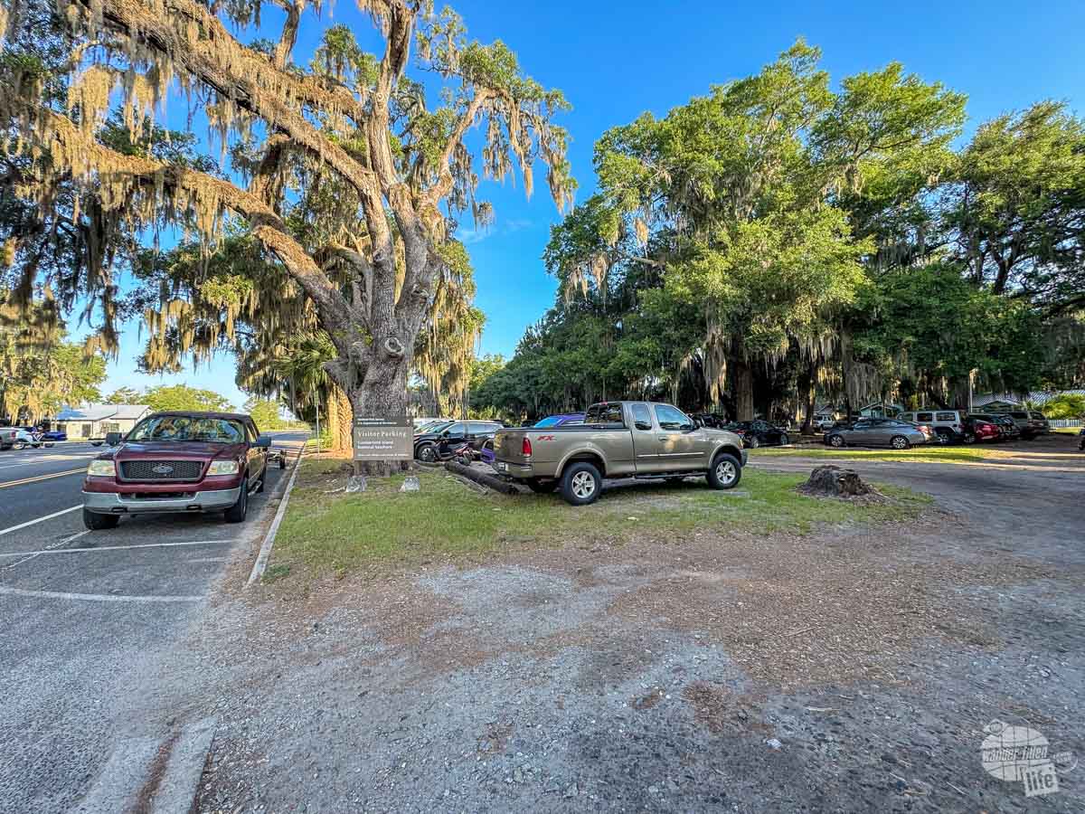 A sandy parking lot with live oaks and several cars parked. There is a sign that says visitor parking on a National Park sign. 