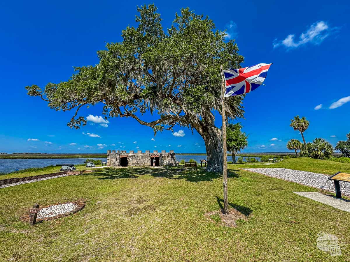 A British flag flies in front of a tree, which overhangs a low stone fortification. There is a river in the distance.