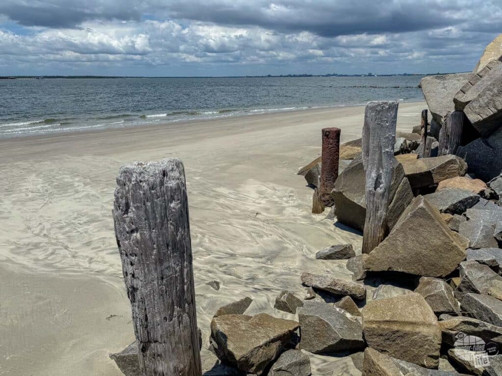 A small beach with rocks and pilings in the foreground. In the far distance is Fort Sumter.