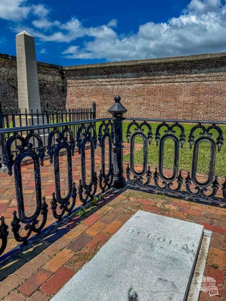 A marble grave marker on a brick walkway with a wrought iron fence surriound it. Beyond the fence is a oblelisk in front of a masonry fort wall.