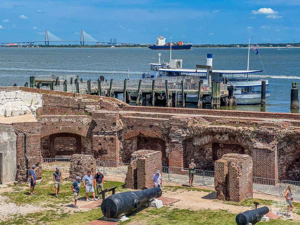 A view of a masonry fort from above with a ferry boat at the dock outside the walls in the harbor. Beyond the boat is harbor traffic and a suspension bridge over Charleston Harbor.