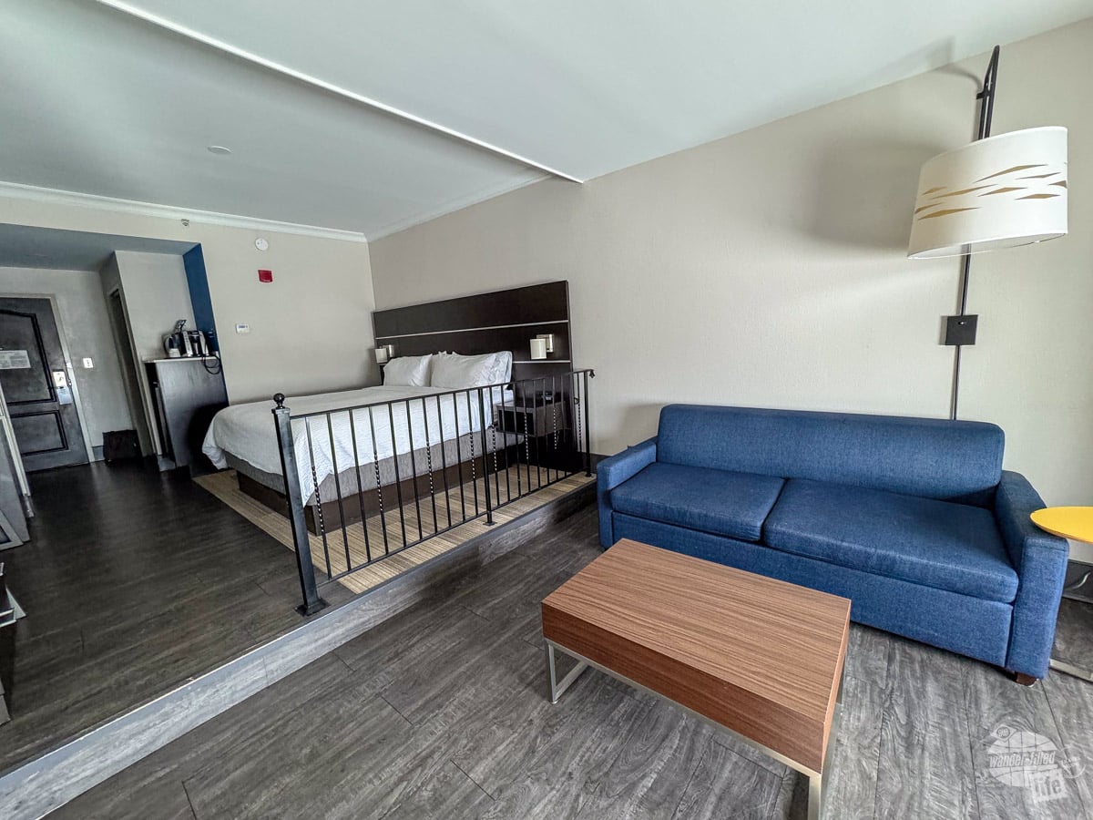 A hotel room with a king bed, night stands, a couch and a coffee table. There is a metal guard rail by the bed and a small step down.