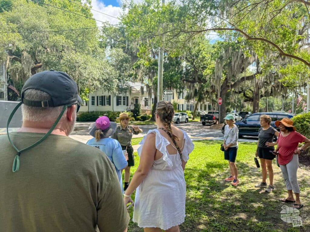 A group of people listening to a park ranger speak in front of a historic home.